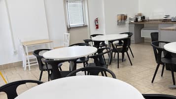Un comedor con mesas y sillas redondas, un extintor en la pared y una ventana con persianas.