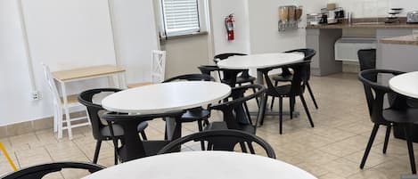 A dining area with round tables and chairs, a fire extinguisher on the wall, and a window with blinds.