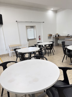 A dining area with round tables and chairs, a fire extinguisher on the wall, and a window with blinds.