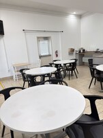 Un comedor con mesas y sillas redondas, un extintor en la pared y una ventana con persianas.