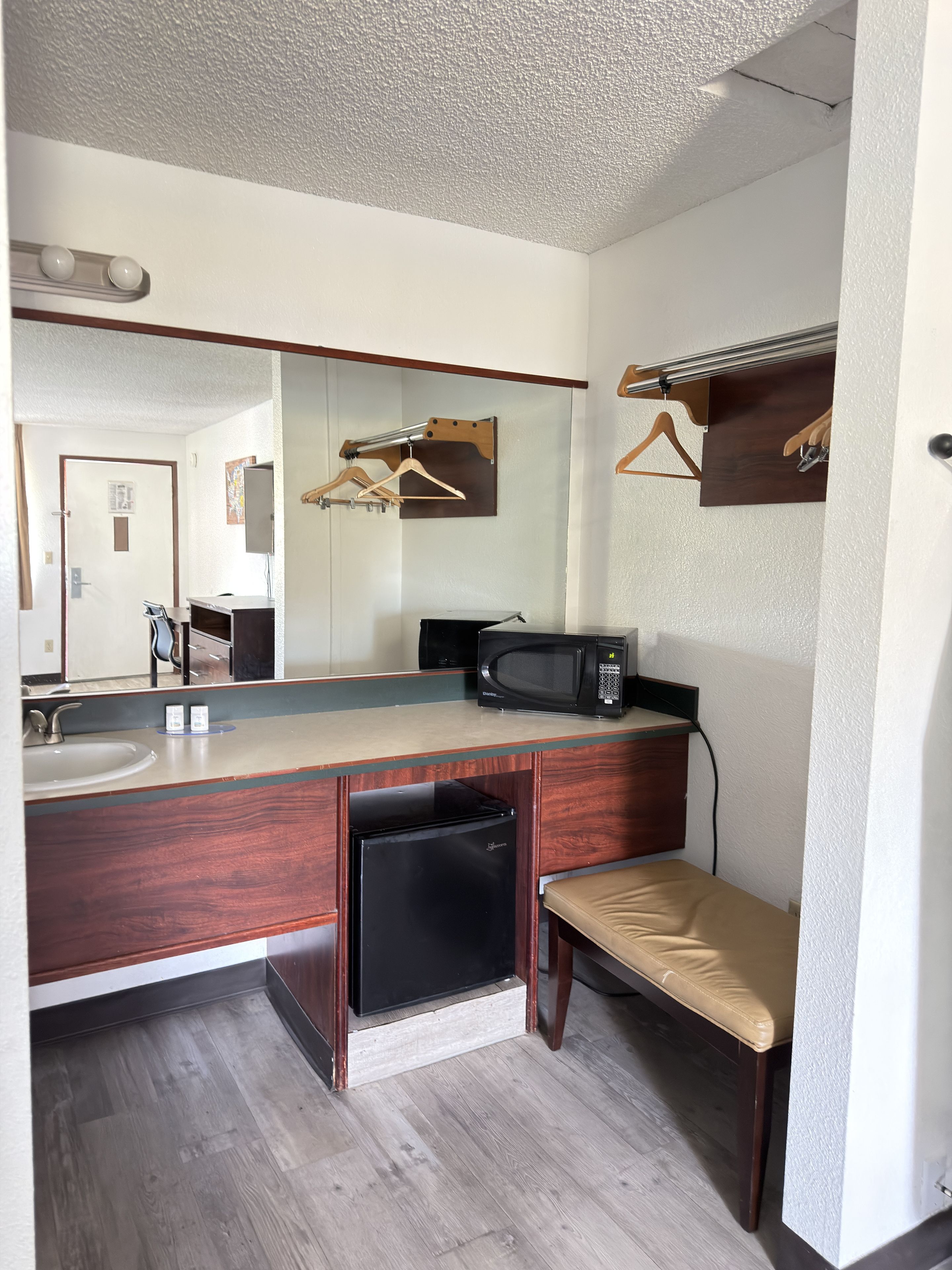 A bathroom with a wooden vanity, a sink, a mirror, a black refrigerator, a microwave, and a bench.
