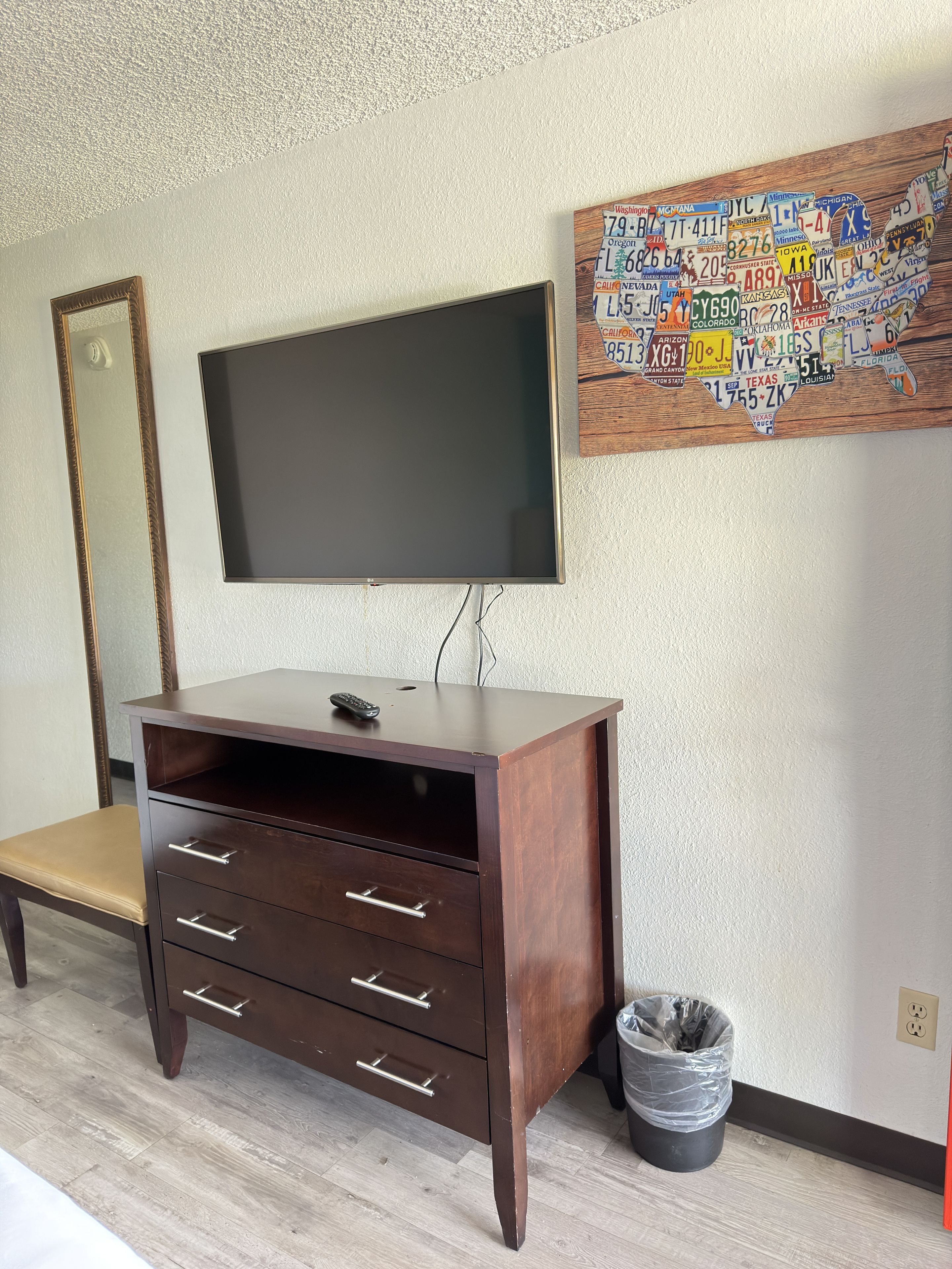 Een kamer met een aan de muur bevestigde televisie, een houten kast met lades, een bankje en een decoratief kunstwerk met nummerborden van een staat.