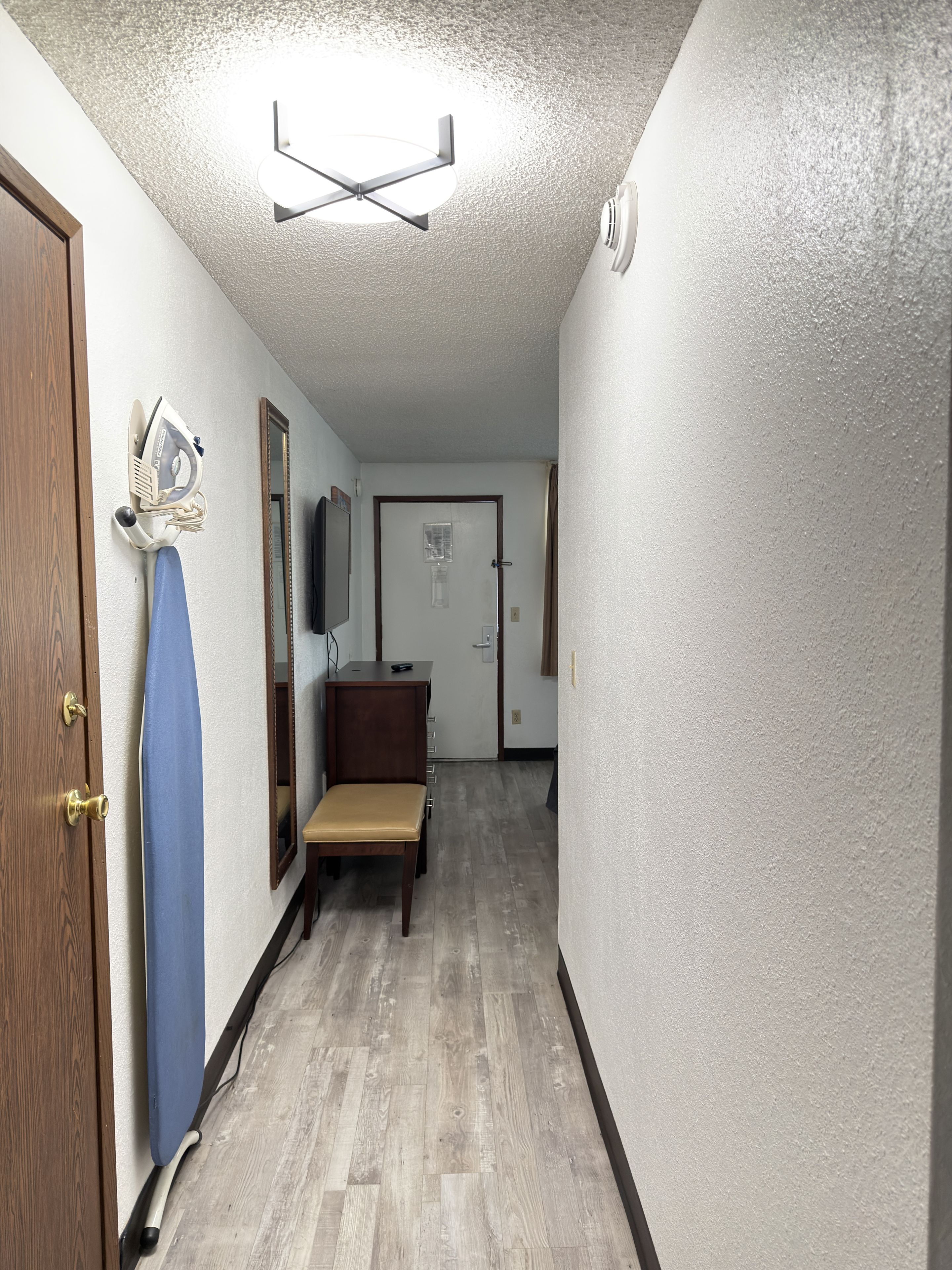 A hallway with a wooden door, a blue ironing board, a small table with a chair, and a mirror on the wall.