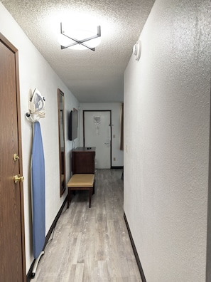 A hallway with a wooden door, a blue ironing board, a small table with a chair, and a mirror on the wall.