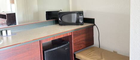 A compact kitchenette with a microwave, a small refrigerator, a bench, and a wooden shelf with hangers.