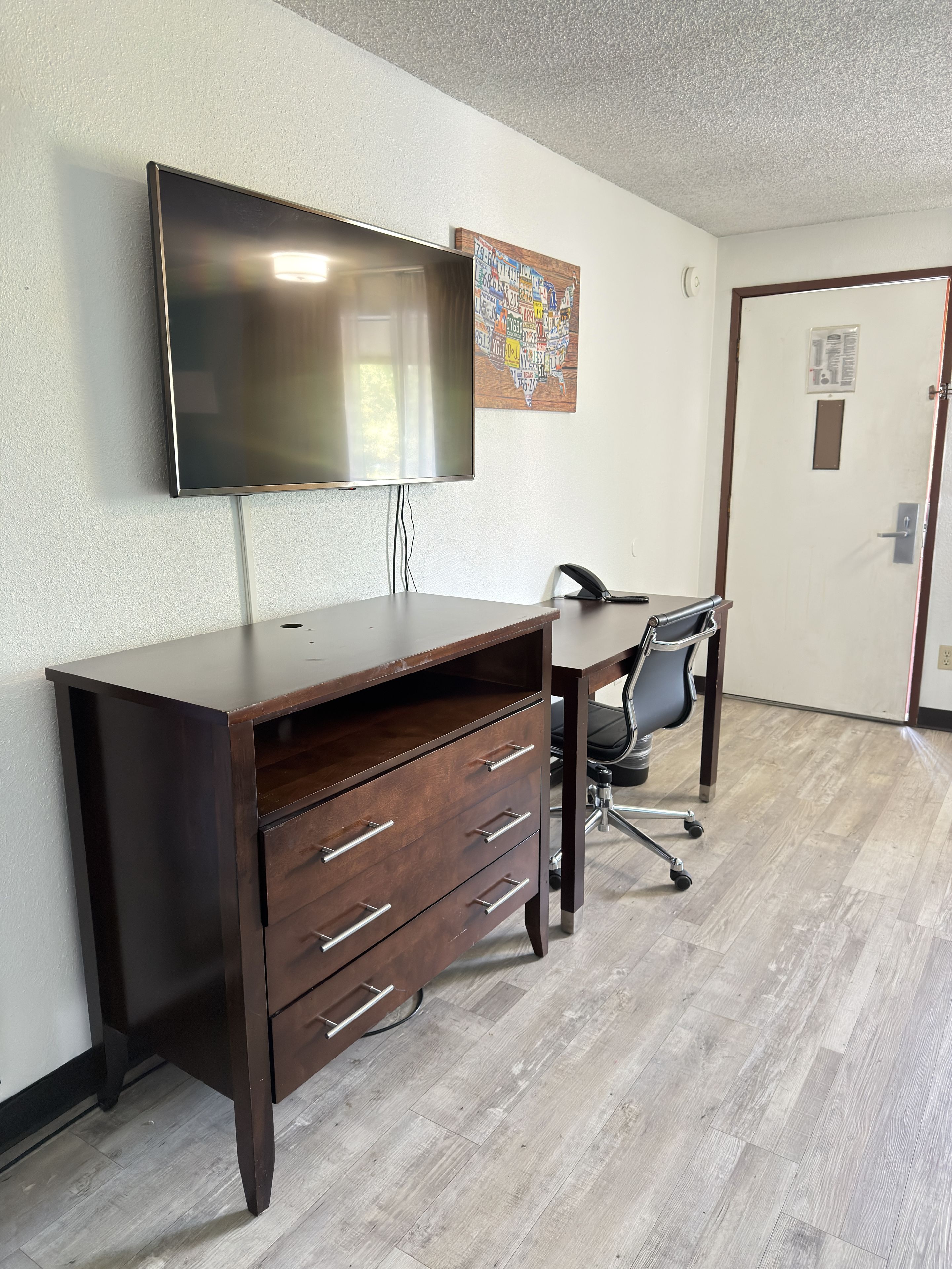 A conference room with a large flat-screen TV mounted on the wall, a wooden desk with drawers, a black office chair, and a door in the background.