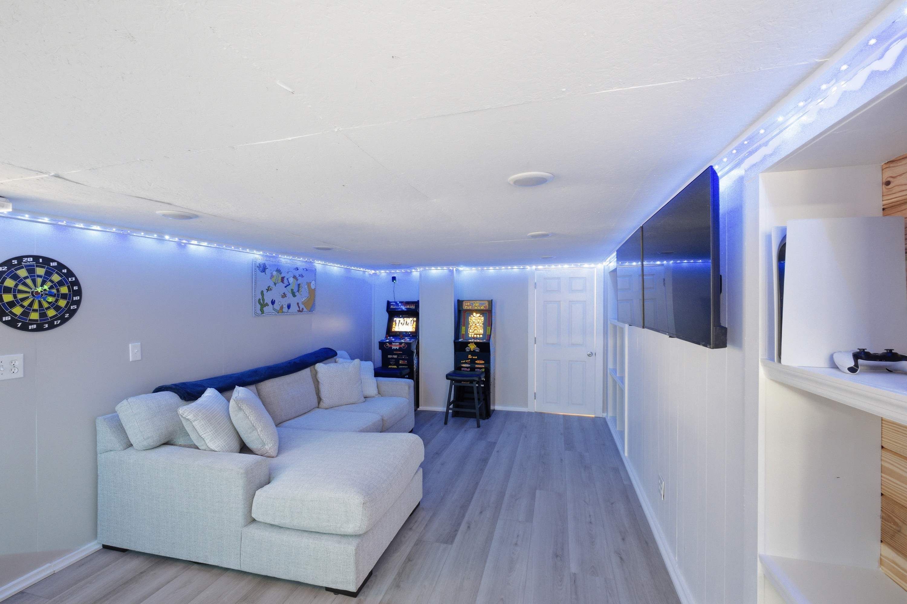 A living room with a sectional sofa, dartboard, arcade games, and a flat-screen TV.