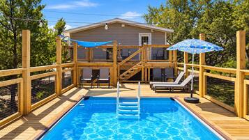 Una piscina con muelle de madera, sillas de descanso y una casa al fondo.