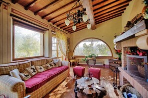 Ein gemütliches Zimmer mit Holzbalkendecke, einem großen Fenster, einem Kamin und einer Sitzecke mit Sofa und Hocker.