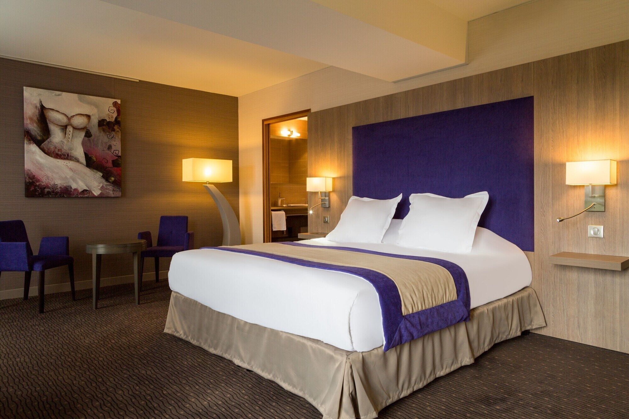 A hotel room with a large bed, two bedside lamps, a desk with a chair, and a painting on the wall.