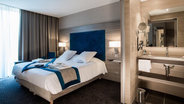 A modern hotel room with a large bed, bedside lamps, a bathroom with a mirror, sink, and towel rack. - Relais Du Silence Domaine La Charpiniere (Saint-Galmier)