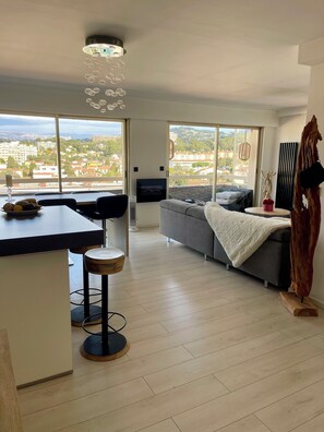 A modern living space with a kitchen area, a sofa, a dining table with chairs, and large windows offering a city view. - Magnificent 3-room apartment (Le Cannet)
