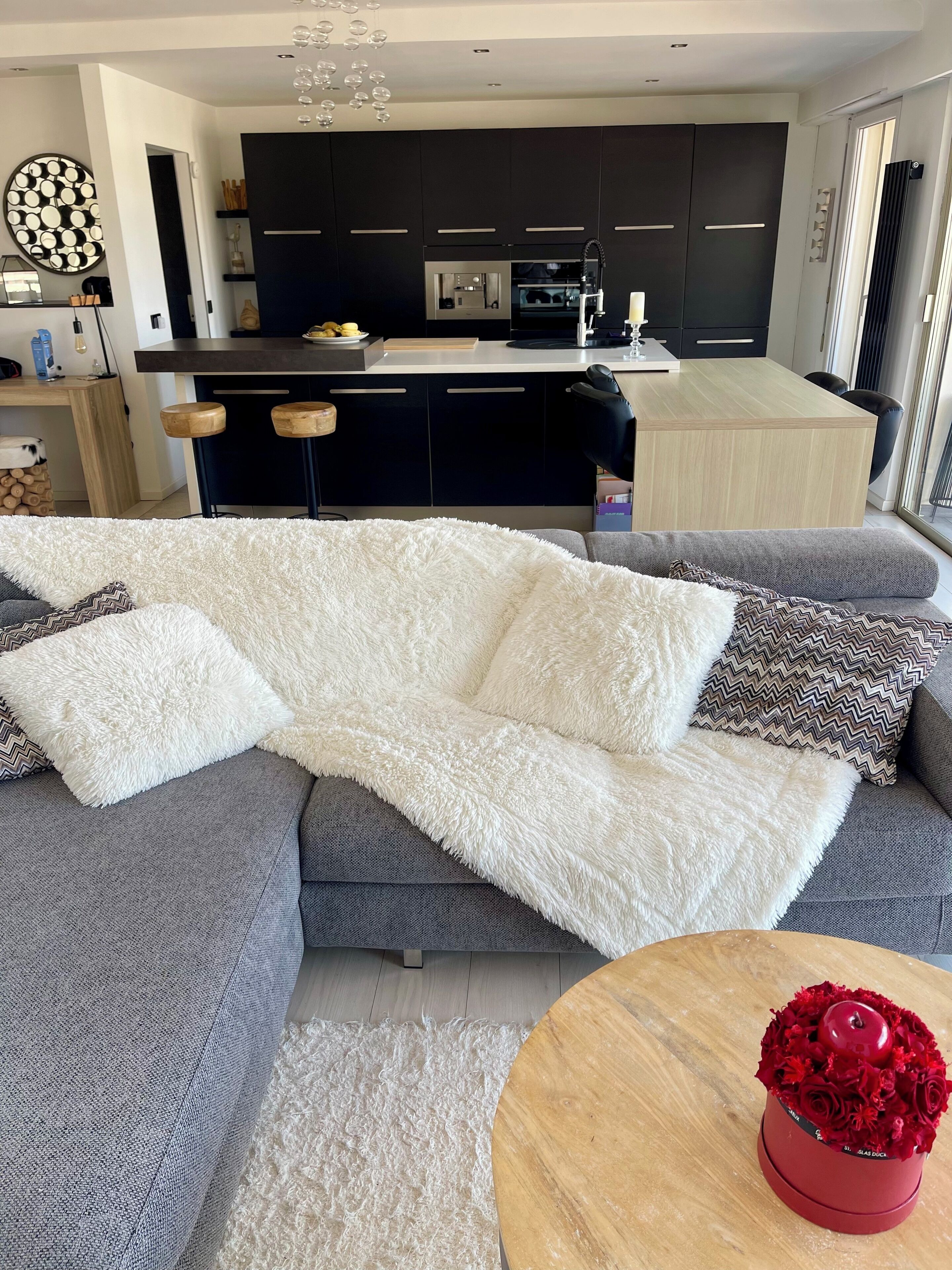Een moderne woonkamer met een grijze bank, een witte zachte plaid en een ronde houten salontafel met een rode decoratie.