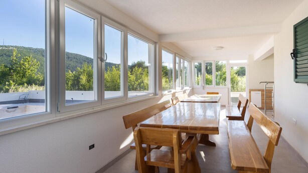 Un espace repas avec une longue table et des bancs, de grandes fenêtres et une vue sur un paysage verdoyant.