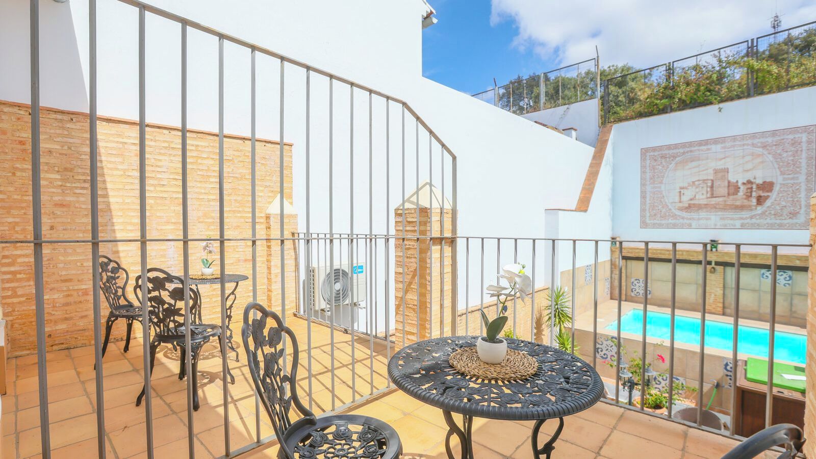 An outdoor patio with metal chairs and a table, a pool in the background, and a tiled wall with a decorative pattern.