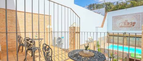An outdoor patio with metal chairs and a table, a pool in the background, and a tiled wall with a decorative pattern.