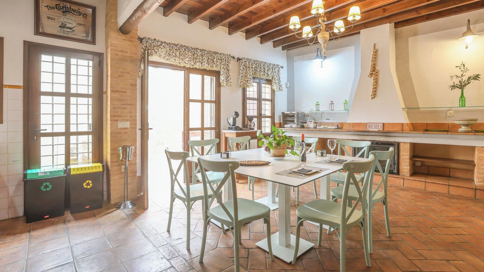 Un coin repas avec une table dressée pour quatre personnes, entourée de chaises, dans une pièce dont le plafond est orné de poutres en bois, avec un espace cuisine en arrière-plan.