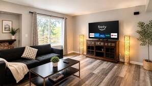 Uma sala de estar moderna com um sofá preto, uma mesa de centro, uma TV na parede exibindo serviços de streaming e uma grande janela com cortinas.