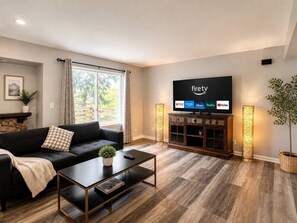 A modern living room with a black sofa, a coffee table, a TV mounted on the wall displaying streaming services, and a large window with curtains. - Insurance & Workers | 5 Beds | Easy S (Grayslake)