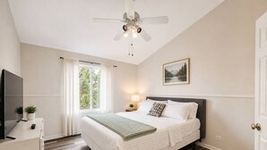 Quarto com cama, ventoinha no teto, televisão, escrivaninha e janela com cortinas.