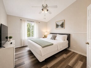 A bedroom with a bed, a ceiling fan, a TV, a desk, and a window with curtains.
