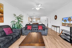 Ein Wohnzimmer mit einer Sofagarnitur, einem Couchtisch, einer TV-Konsole, einem Deckenventilator und einer großen Pflanze.