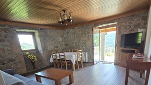 Ein gemütlicher Wohnbereich mit Steinwand, Holzbalkendecke, einer Couch, einem Couchtisch, einem Fernseher und einem Esstisch mit Stühlen.
