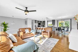 A modern living room with a sofa, armchair, and coffee table, a kitchen in the background, and a dining area with chairs. - Spacious 4BR Home Near O’Hare | Mid-Term Friendly (Bensenville)