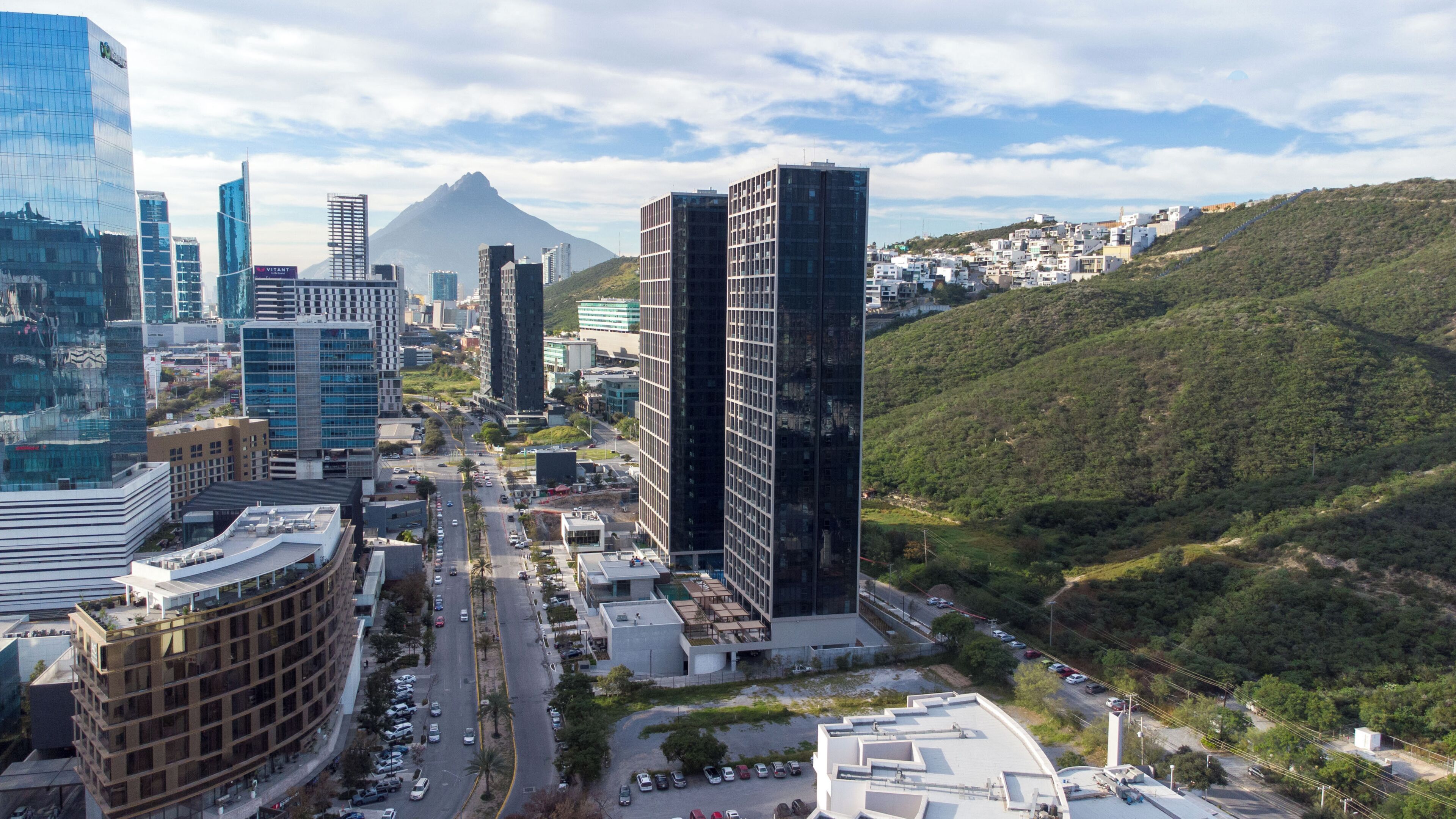 Un paisaje urbano con edificios modernos, una montaña destacada al fondo y un terreno montañoso.