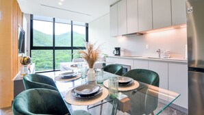 Un espace de salle à manger moderne avec une table en verre, des chaises vertes et une vue sur des collines verdoyantes, visible à travers de grandes fenêtres.
