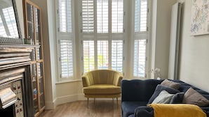 A living room with a dark blue sofa, a yellow armchair, a fireplace, and large windows with shutters. - Modern 3-Bed Home | Walk to Town & Metro | Sleeps 7 (Altrincham)