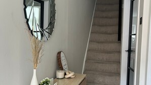 A hallway with a wooden bench, a wall-mounted mirror, a small potted plant, and a staircase in the background. - Modern 3-Bed Home | Walk to Town & Metro | Sleeps 7 (Altrincham)