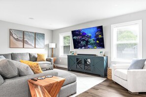 A living room with a grey sofa, a white armchair, a wooden coffee table, a TV, and framed artwork on the wall.