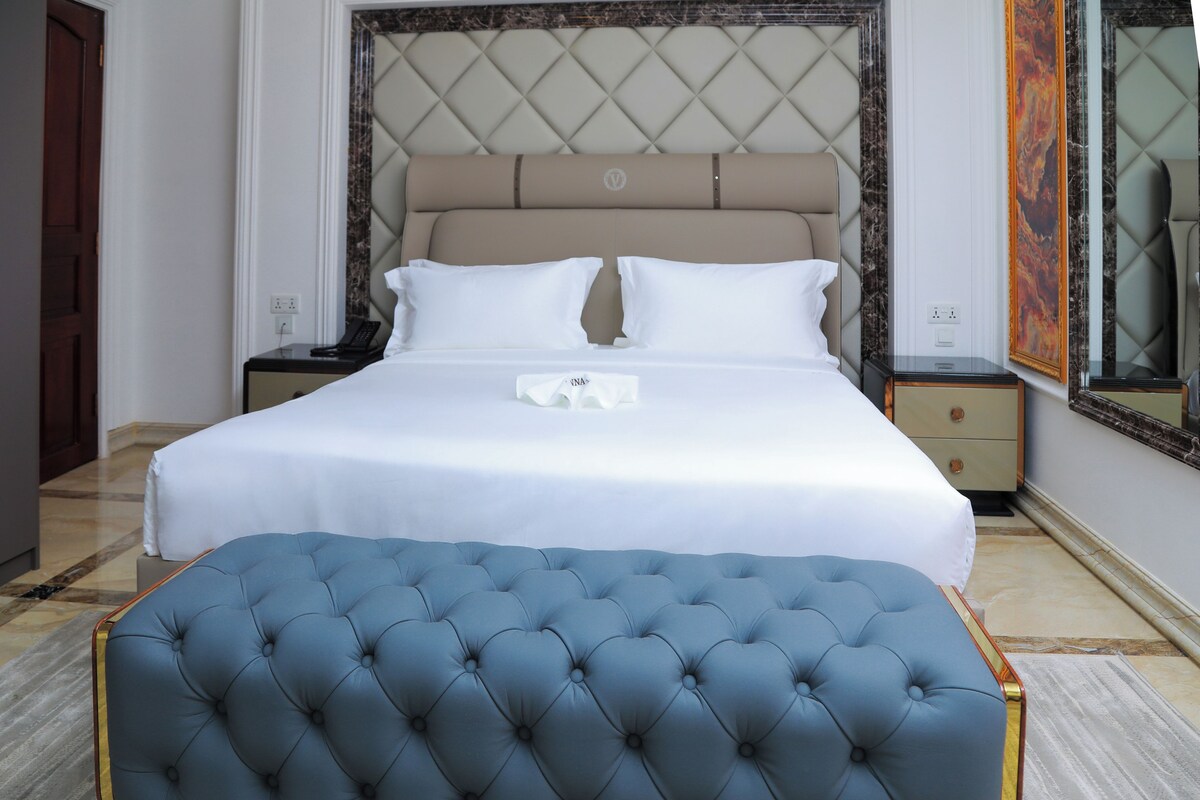 A neatly made bed with white linens, a tufted blue ottoman, and a headboard with a diamond pattern.