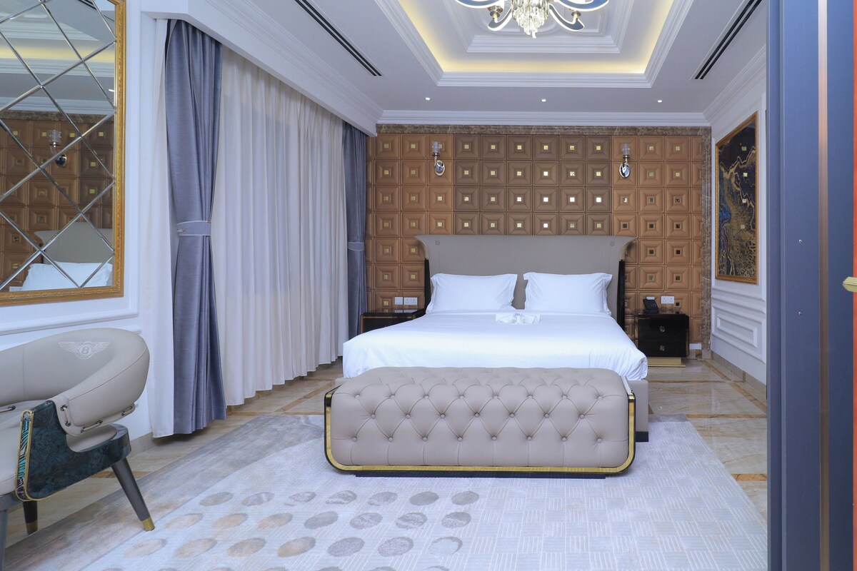 A bedroom with a large bed, a tufted bench, a nightstand, and a chandelier.