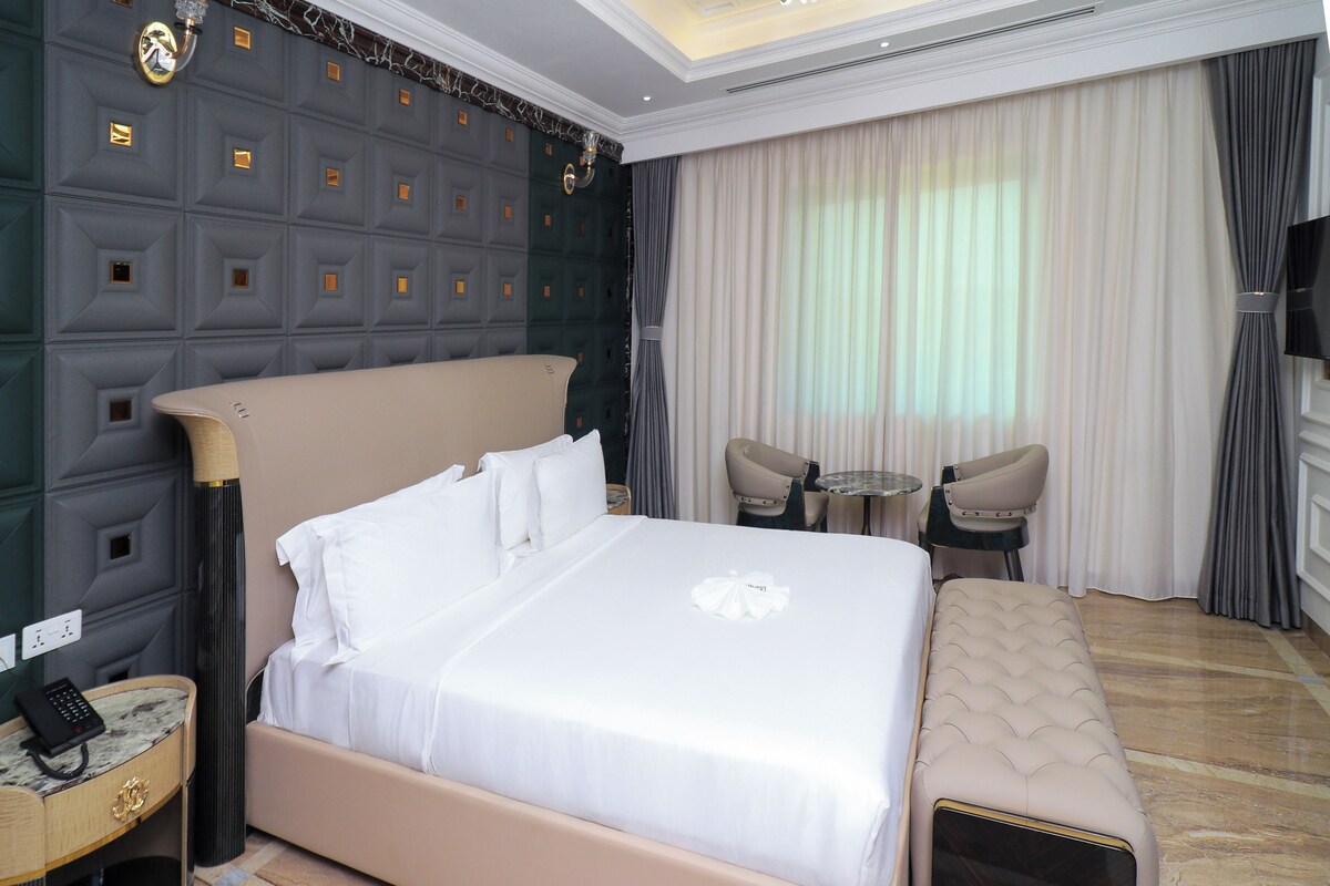 A hotel room with a large bed, a small round table, two chairs, a bench, a TV, and a wall with a geometric pattern.