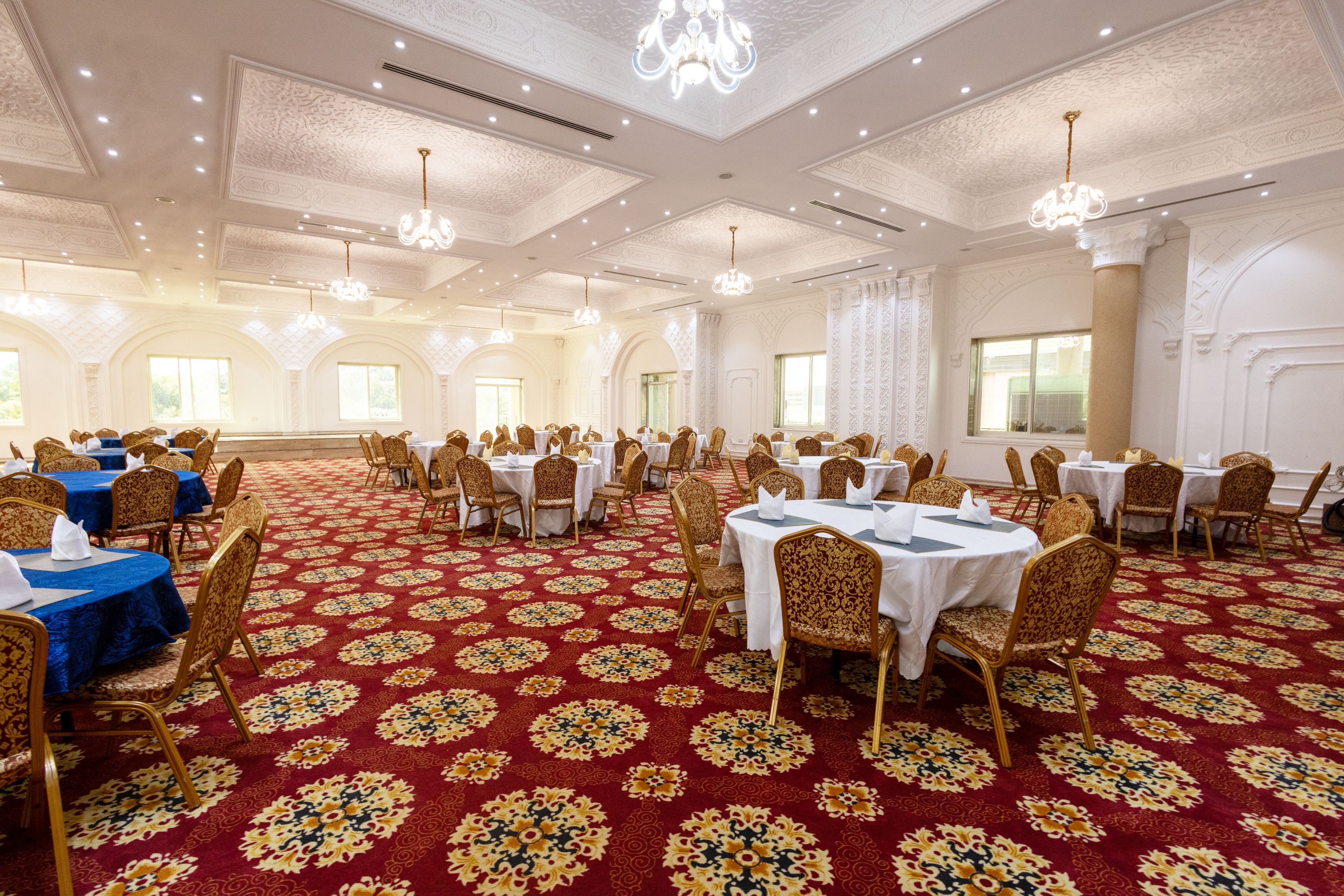 A banquet hall with round tables and chairs, ornate ceiling, and large windows.