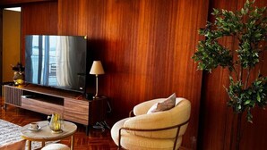 A modern hotel room with a wooden floor, a large flat-screen TV, a sofa, a chair, a lamp, and a potted plant. - Skyline Sea Luxe (Thessaloniki)