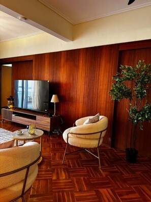 Ein modernes Hotelzimmer mit Holzboden, einem großen Flachbildfernseher, einer Couch, einem Sessel, einer Lampe und einem Topf mit Pflanze.
