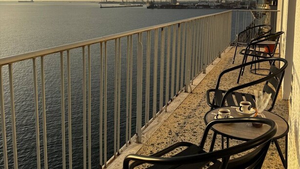 A balcony with a railing overlooking a body of water, two chairs, and a table with cups and a muffin. - Skyline Sea Luxe (Thessaloniki)