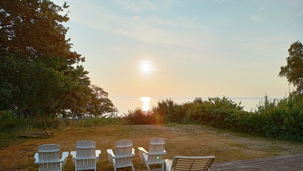 A wooden deck with white chairs and a lounge chair facing a sunset over water. - Holiday home with sea view in Sandkaas (Allinge-Sandvig)