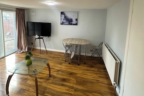 Uma sala de estar com uma mesa de centro de vidro, uma mesa dobrável, uma televisão de tela plana e um radiador.