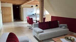Ein modernes Wohnzimmer mit einem grauen Sofa, roten Kissen, einem Holztisch und einem Essbereich im Hintergrund.