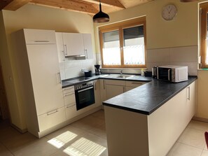 A kitchen with white cabinets, a black countertop, a sink, a stove, a microwave, and a window with blinds. - Beautiful vacation apartment, idyllic landscape, near Rothenburg, ob der Tauber (74572 Blaufelden)