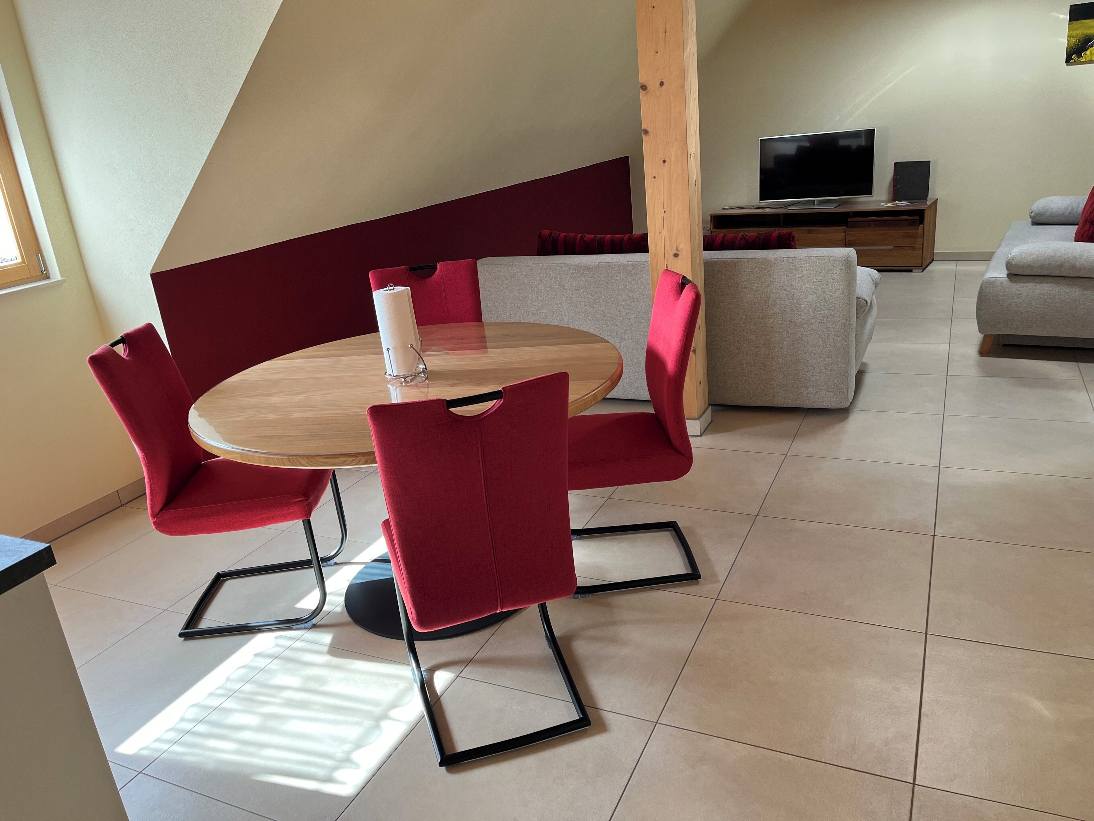 Uma sala de estar moderna com uma mesa de madeira redonda, cadeiras estofadas em vermelho, um sofá cinza e uma televisão sobre um móvel de madeira.