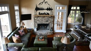 A cozy living room with a stone fireplace, a sofa set, a dining table, and a TV mounted on the wall. - Lake-Front Family Home in Big Canoe (Jasper)