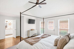 A bedroom with a four-poster bed, a ceiling fan, a television on the wall, and a view of a brick building through the windows. - Renovated Retreat w/ Game Room & Free Parking (Montgomery)