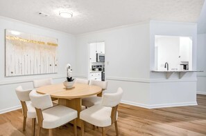A dining area with a round wooden table and six upholstered chairs, a white wall with a large abstract painting, and a kitchen visible through an open doorway. - Renovated Retreat w/ Game Room & Free Parking (Montgomery)