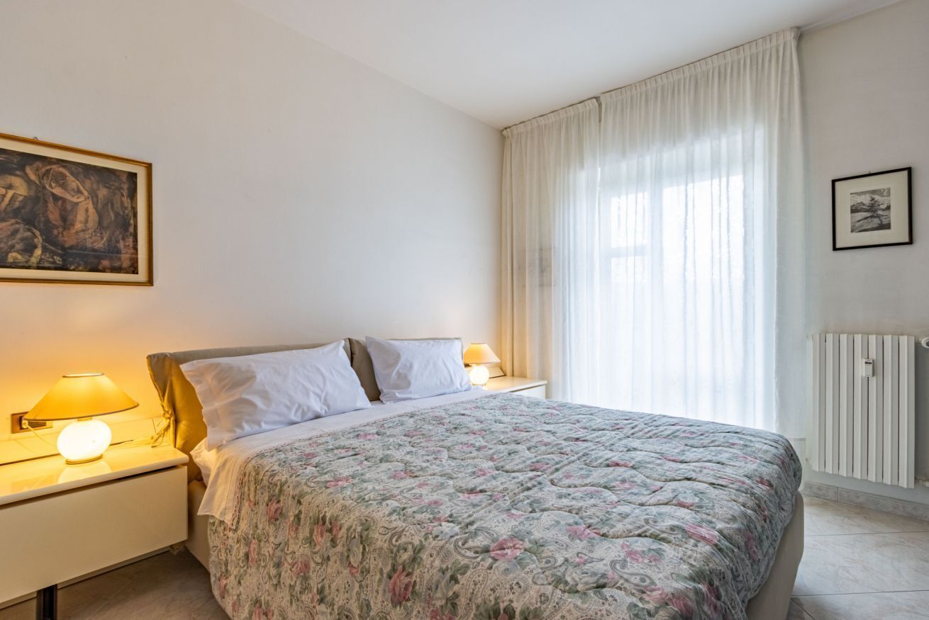 Een slaapkamer met een bed, nachtkastjes, lampen, een schilderij, een raam met gordijnen en een radiator.