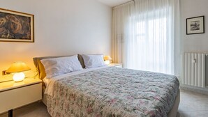 Une chambre à coucher avec un lit, des tables de chevet, des lampes, un tableau, une fenêtre avec des rideaux et un radiateur.